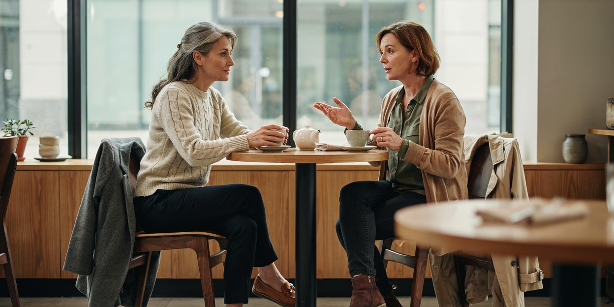 Two women in their fifties talking over coffee at a cafe table by a window