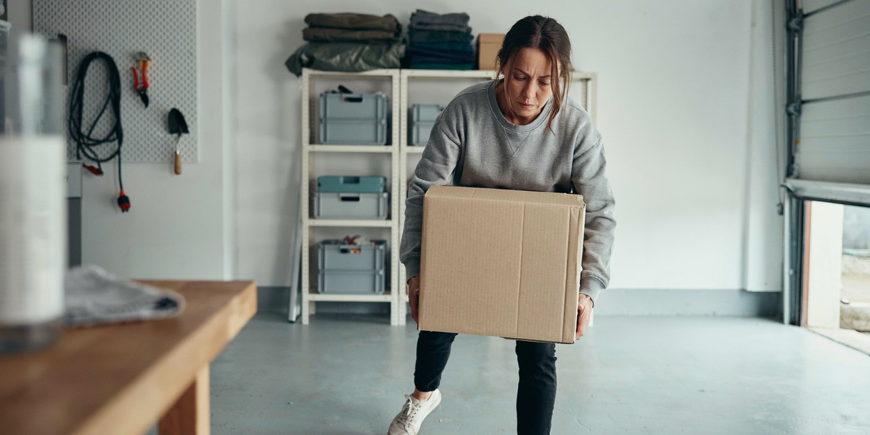 Woman in her forties lifting a heavy cardboard box down from a shelf in a garage